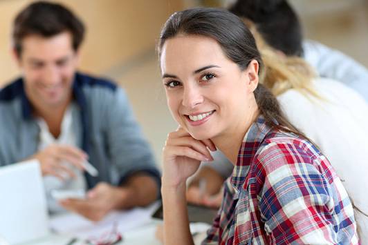 Quelle: Fotolia_57985017_XXL Gruppe Student*innen am Tisch, im Vordergrund junge Frau mit Blick zur/m Betrachter*in